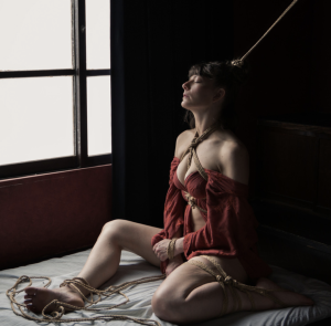 Kissmedeadlydoll seated by a window in a self-tied pose including hair rope attached to an upline